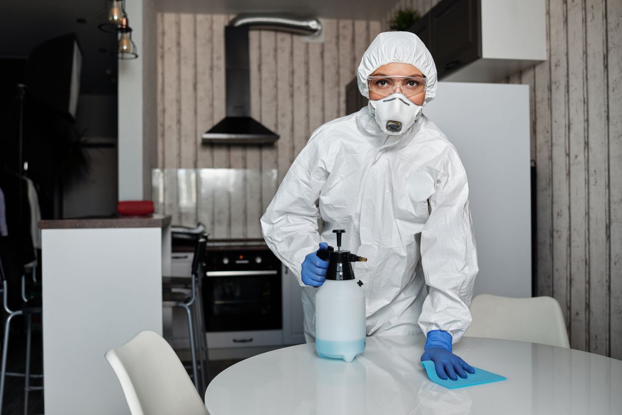 A cleaner in full protective gear disinfects a table indoors, ensuring a sterile environment.