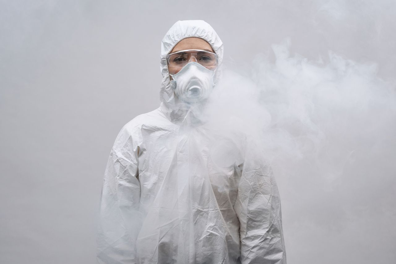 Individual in protective suit and mask standing in smoke on white background.