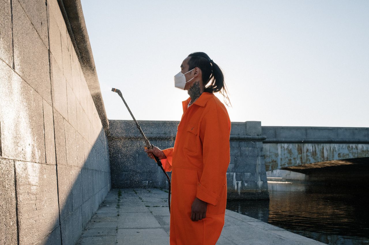 Man in orange jumpsuit and mask cleaning an outdoor urban wall. Bright daylight setting.